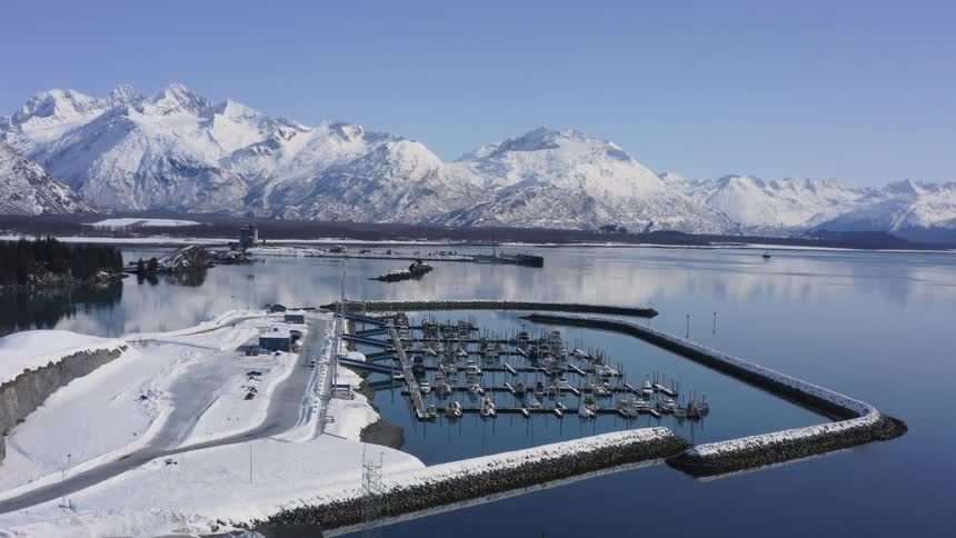 A beautiful aerial shot slowly pulls back to reveal a snowy harbor in Alaska, with boats docked in the calm water against a backdrop of majestic mountains