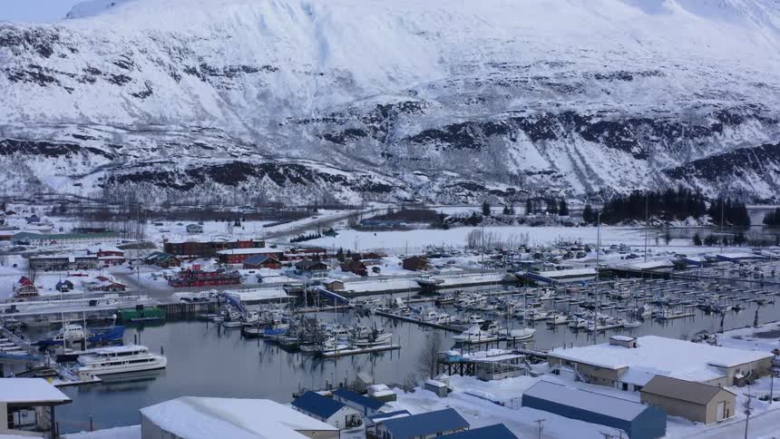 Slow aerial push-in shot of a snowy fishing harbor in a small Alaskan town nestled at the base of a large mountain