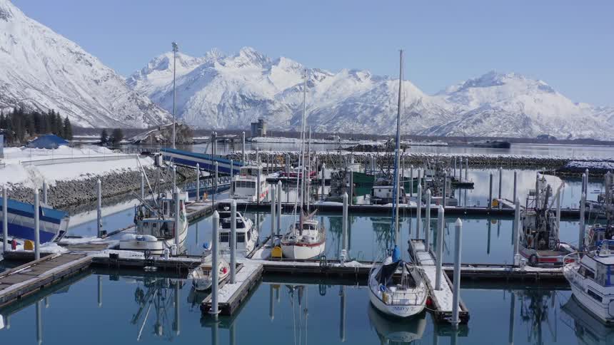 A slow forward aerial reveals boats docked in a tranquil, snowy Alaskan harbor with majestic mountains in the background