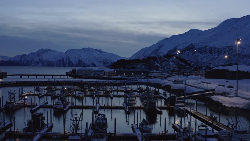 A serene view of a snowy harbor with docked boats and glowing lights against a backdrop of mountains at twilight