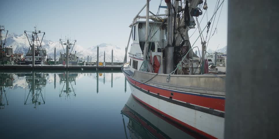 A fishing boat sits peacefully in a calm harbor, its reflection mirrored on the glassy water against a backdrop of snowy Alaskan mountains