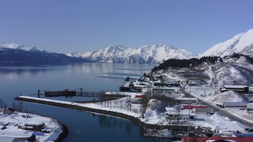 A slow aerial push-in shot of a snowy coastal town with a harbor, surrounded by majestic mountains and a calm bay on a clear winter day