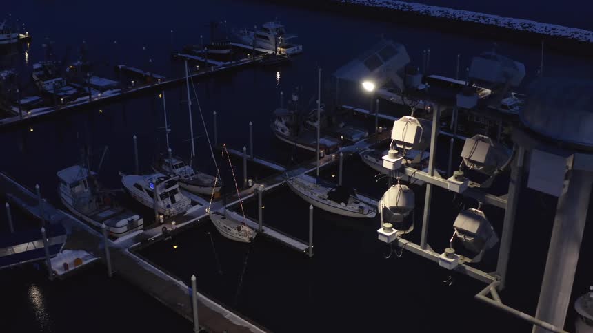 Flying past a large light pole at night to reveal a quiet, snow-covered harbor filled with docked fishing boats and sailboats