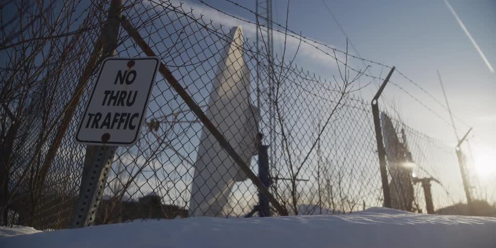 A low-angle shot of a 'No Thru Traffic' sign on a snowy chain-link fence with a large antenna in the background at sunrise