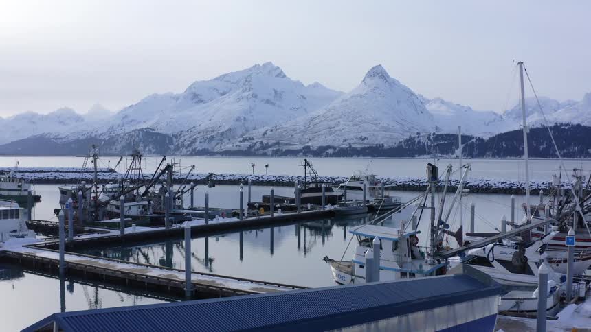 A slow push-in shot reveals a tranquil fishing harbor in winter, with boats docked in calm water against a backdrop of majestic snow-covered mountains