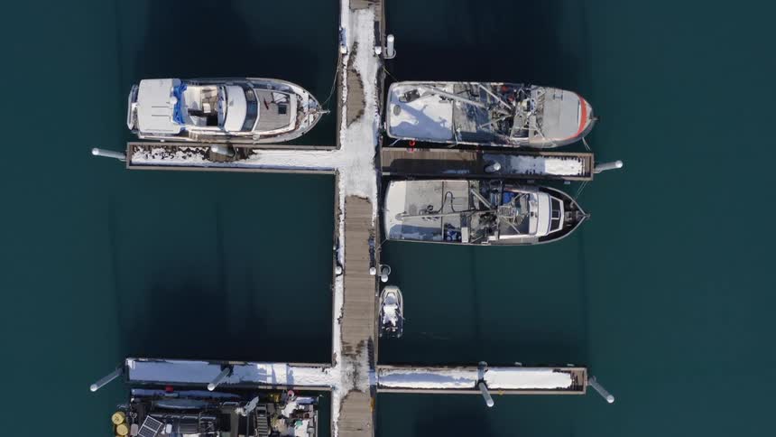 A serene top-down aerial view slowly ascends over a snow-covered marina where various boats are peacefully docked in the calm winter waters