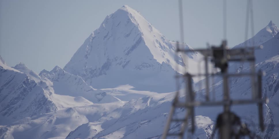 A majestic snow-covered mountain peak stands tall against a clear sky, viewed from behind a blurred ski lift