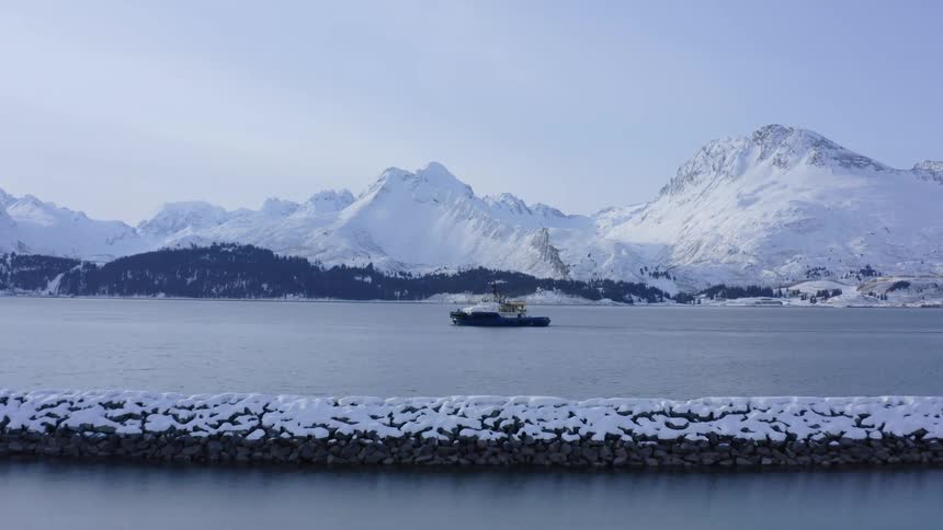 An aerial shot moves from a quiet winter harbor to a tugboat cruising in a bay, with a backdrop of majestic snowy mountains