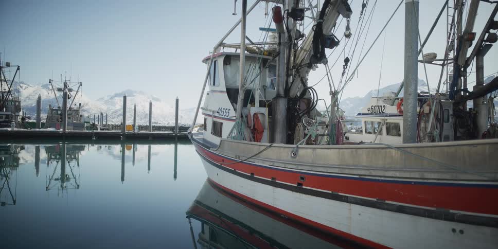 A fishing boat rests in the calm, reflective waters of a harbor with snow-covered mountains in the background