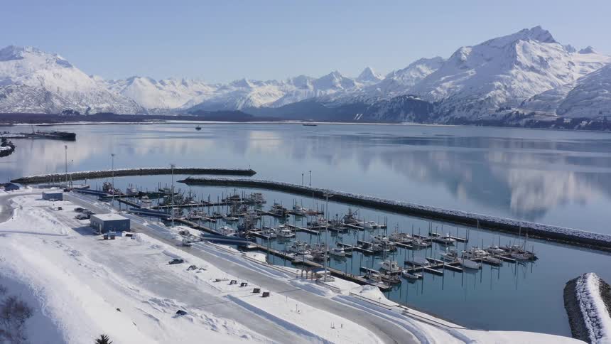 A stunning aerial view captures a tranquil, snow-covered harbor filled with docked boats, set against a backdrop of majestic, snow-capped mountains