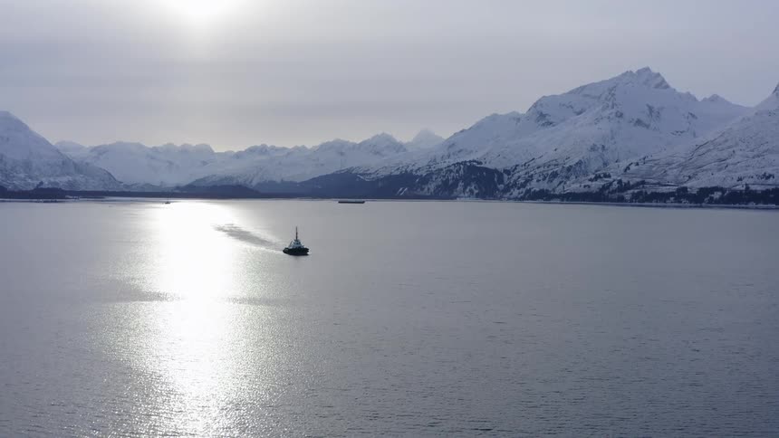A lone tugboat cruises through a vast, icy bay, flanked by towering snow-covered mountains as the sun reflects on the calm water
