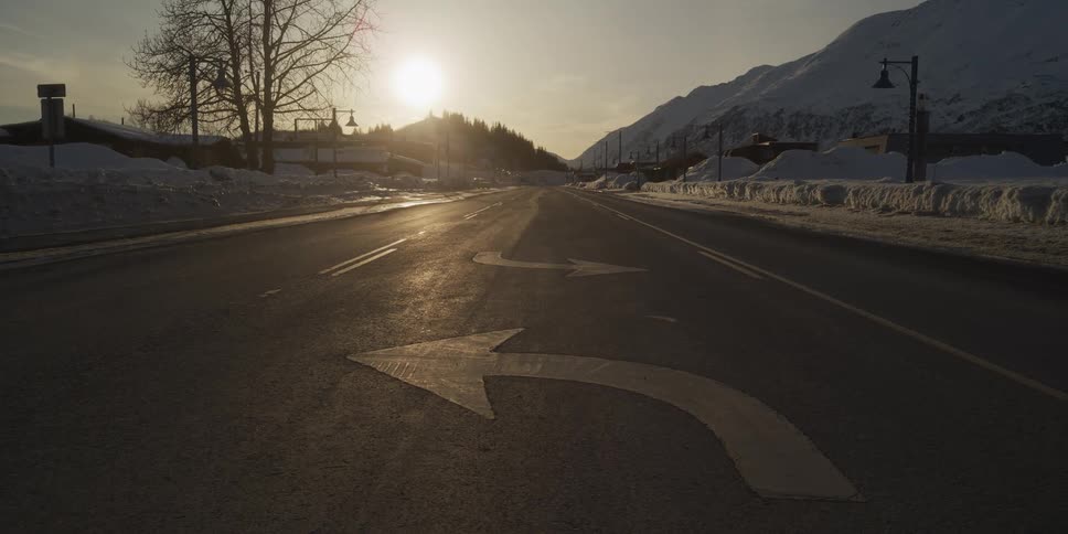 A low-angle view of a quiet, snowy mountain road with turn arrows painted on the asphalt as the sun sets in the distance