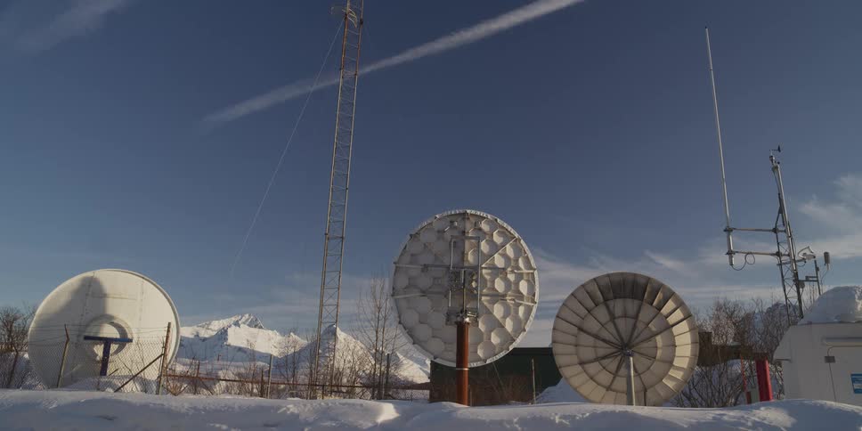 A wide shot shows a remote communication station with satellite dishes and antennas in a snow-covered landscape with mountains in the background