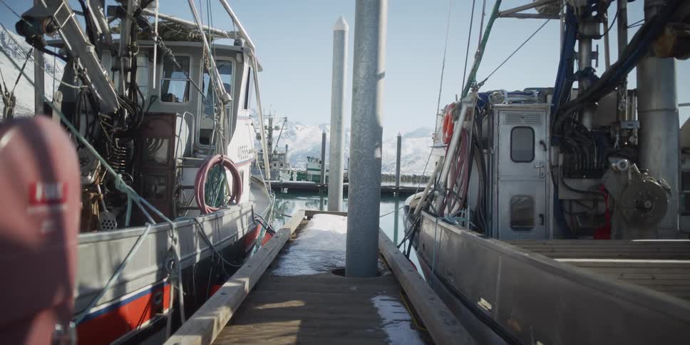 A tranquil scene of fishing boats docked along a snowy pier in a sunny harbor with mountains in the background