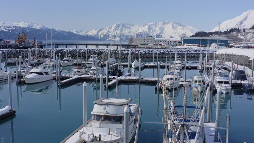 A serene aerial shot slowly pushes over a tranquil, snow-covered boat harbor with majestic mountains in the background during winter