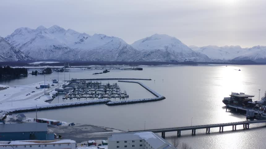 An aerial view slowly zooms in on a tranquil, snow-covered harbor in Valdez, Alaska, with majestic mountains in the background