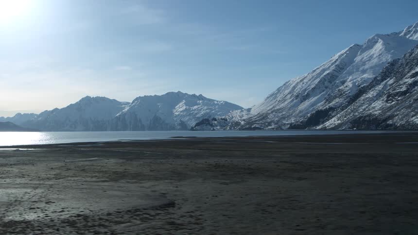 A vast, sunlit winter landscape reveals a dark, frozen shoreline and a tranquil lake surrounded by towering snow-capped mountains
