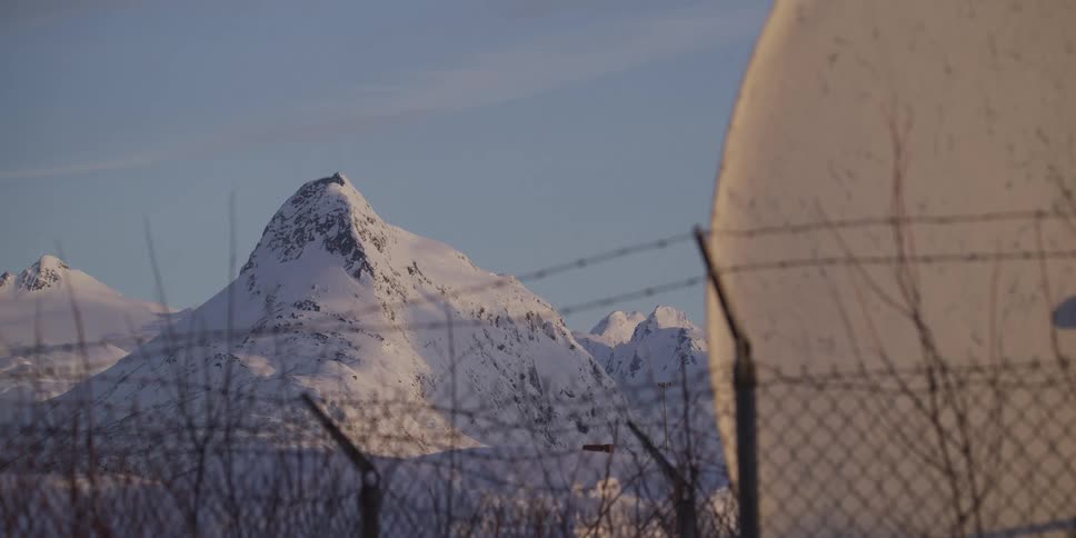 A snow-covered mountain peak stands in the distance behind a satellite dish and a barbed wire fence