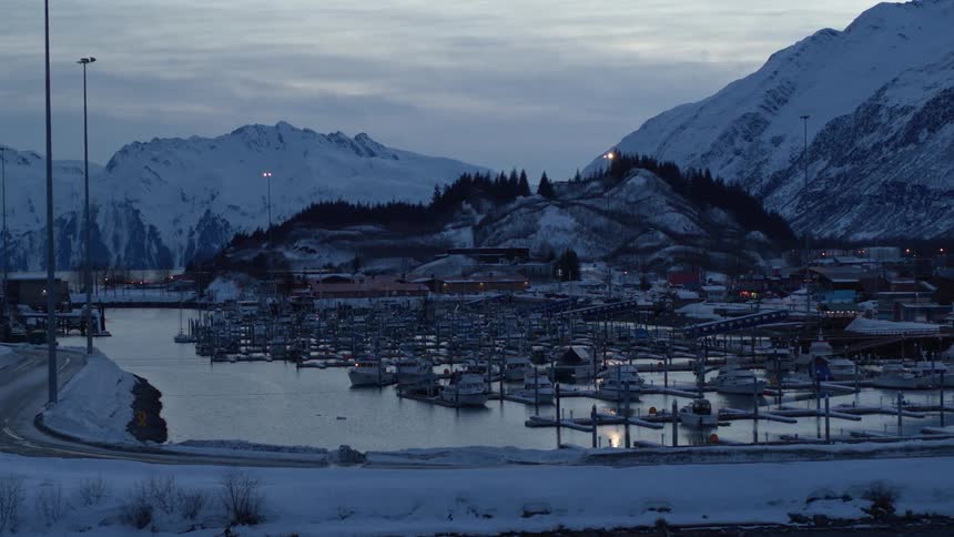 A wide shot of a peaceful harbor filled with boats in a snowy mountain town at dusk