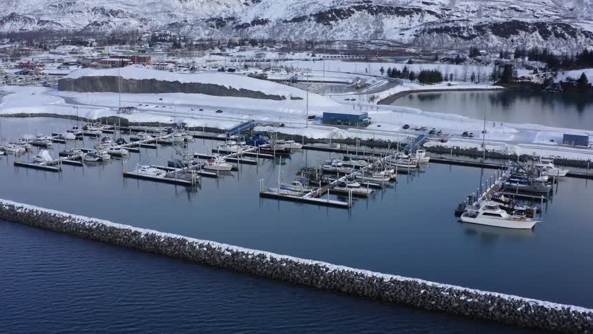 An aerial view slowly pushing in on a snowy boat harbor in winter, with snow-covered mountains in the background