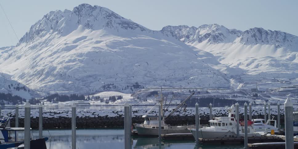 Boats sit docked in a tranquil harbor with a stunning backdrop of snow-covered mountains on a clear winter day