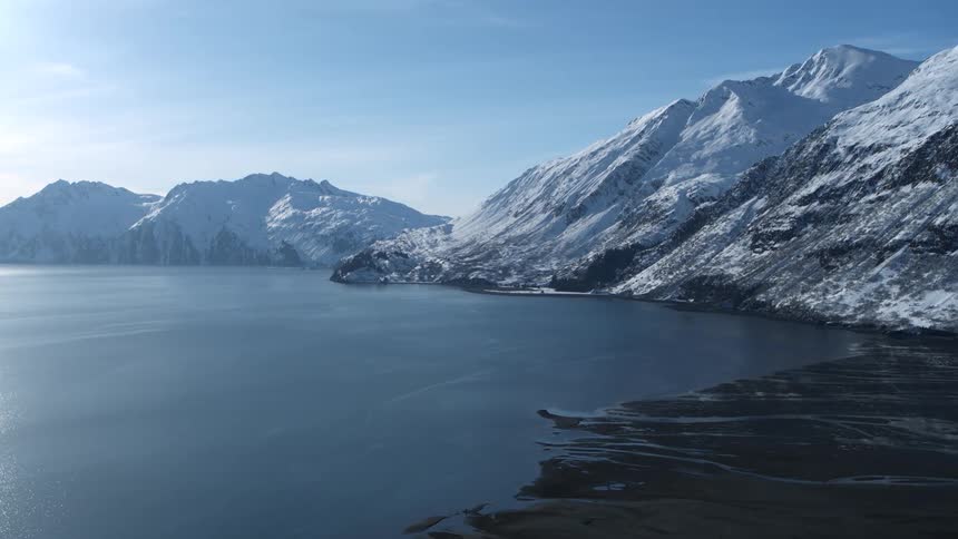 A stunning aerial view shows a vast, calm body of water surrounded by magnificent snow-covered mountains on a clear, sunny day