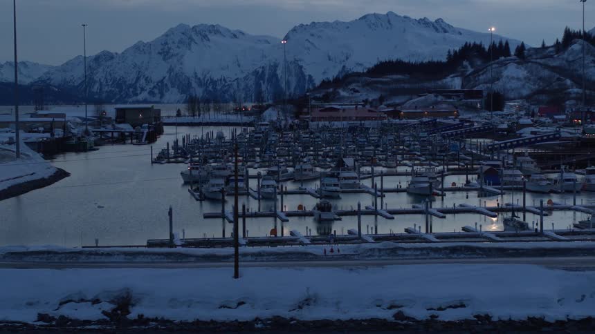 A slow dolly shot of a peaceful, snow-covered harbor in a remote town, with boats docked against a backdrop of majestic mountains at twilight