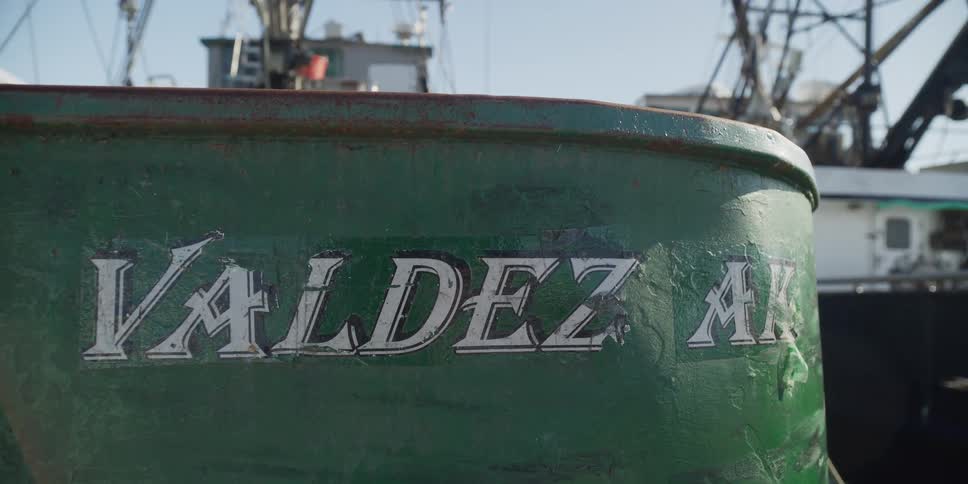 Close-up of a weathered 'Valdez, AK' sticker on the hull of a green fishing boat in a sunny harbor