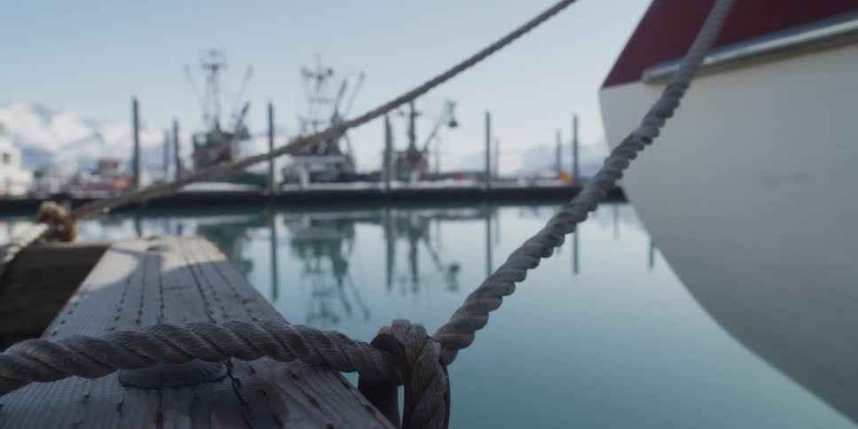 A boat is secured to a dock by a rope, with a fishing harbor and snowy mountains softly focused in the background