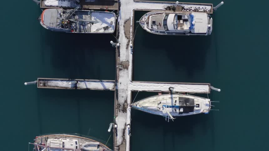 A slow ascending aerial top-down view reveals snow-covered boats moored at a tranquil marina during a cold winter day