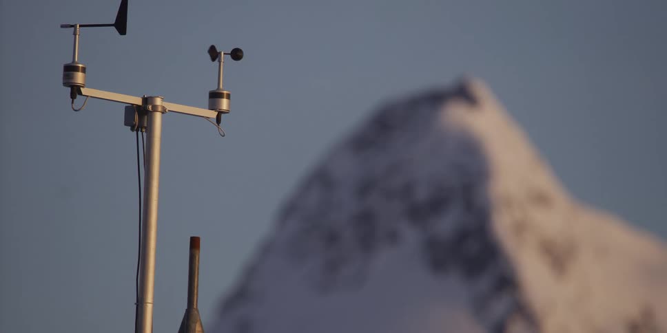 A weather station's anemometer spins in the wind to measure its speed, with a large, snow-covered mountain peak blurred in the background