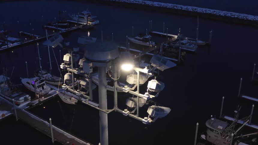 A slow aerial pan reveals a quiet, snow-covered boat harbor illuminated by a large light fixture during the blue hour