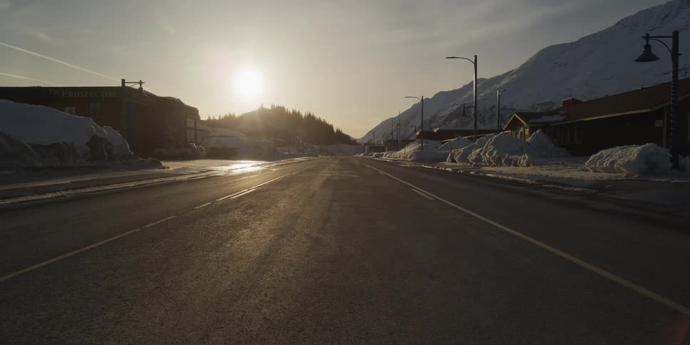 Golden hour sunlight shines down an empty street in a small town surrounded by snowy mountains in winter