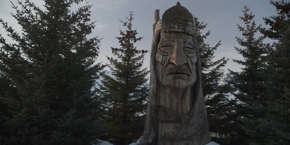 An intricately carved wooden statue of a Native American chief stands among evergreen trees in a snowy winter landscape