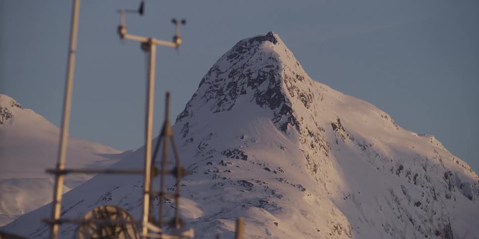 A snow-covered mountain peak stands majestically against a clear morning sky, with a weather station in the soft-focused foreground