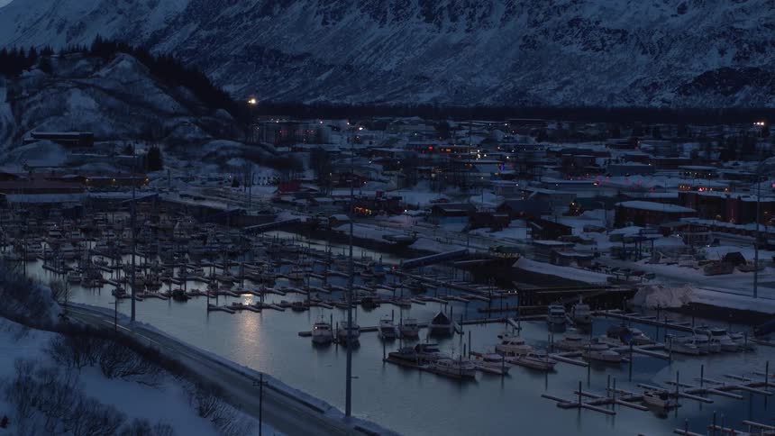 A tranquil evening settles over a snowy harbor town with docked boats and glowing lights at the foot of a mountain