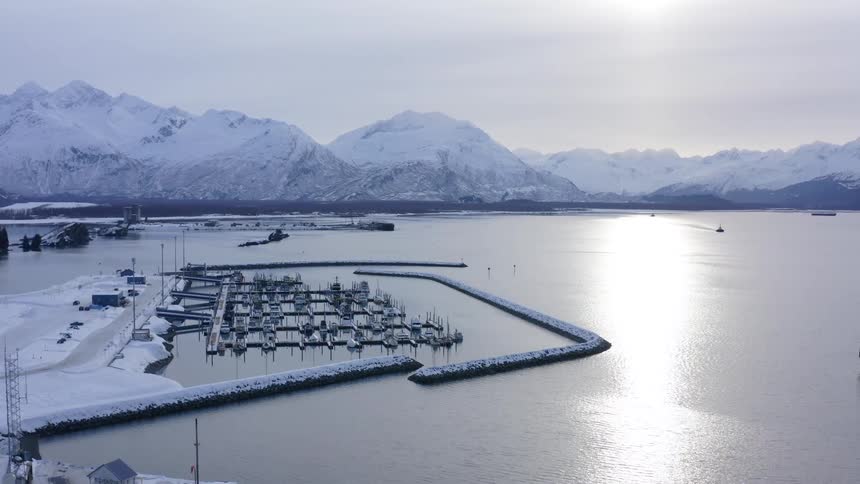 A bright winter sun reflects on the calm waters of a harbor nestled amongst snowy Alaskan mountains