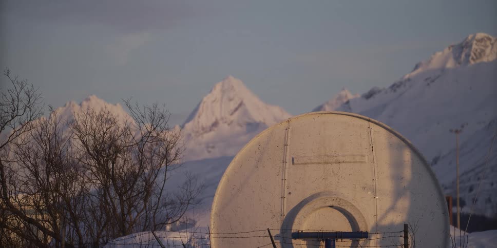 A snowy basketball court with a weathered backboard sits in front of a stunning mountain range during a winter sunset