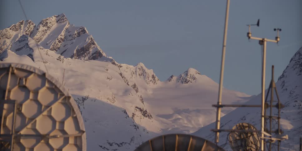 A communication array and weather station are set against a backdrop of majestic, snow-covered mountains