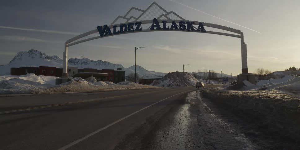 A white truck drives under the Valdez, Alaska welcome sign with snowy mountains in the background at sunset