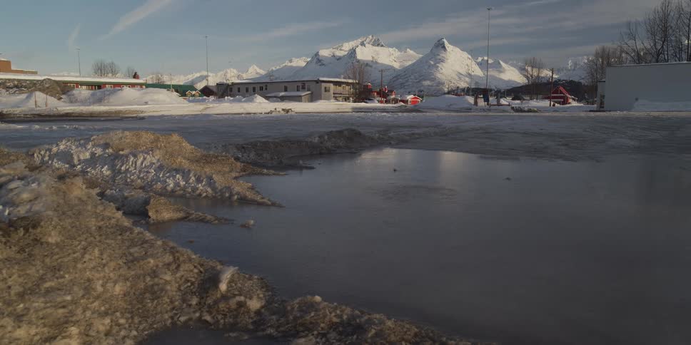 Slow pan right across a frozen puddle in a snowy parking lot with majestic mountains in the distance
