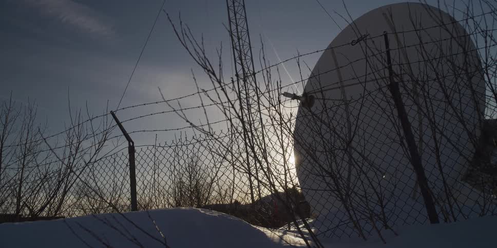 A satellite dish sits behind a barbed wire security fence in a snowy landscape as the sun sets