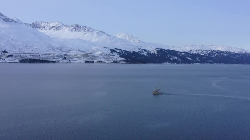 Aerial view of a solitary fishing boat sailing through a calm bay against a backdrop of snow-covered Alaskan mountains