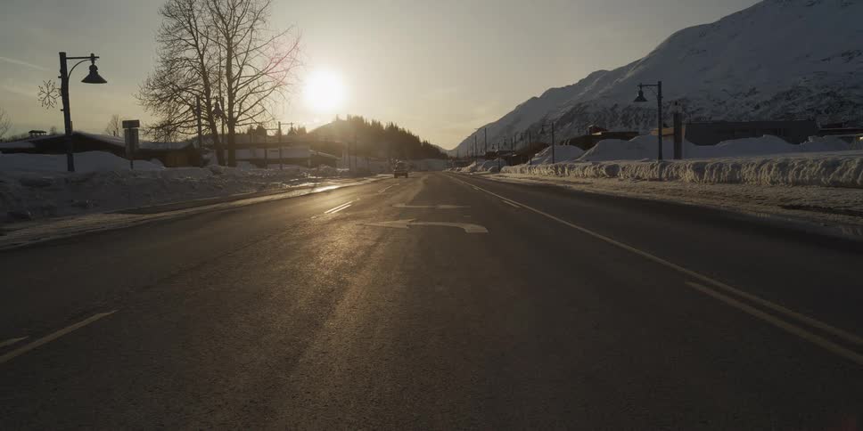 An SUV drives down a quiet, snowy road in a mountain town as the golden sun sets