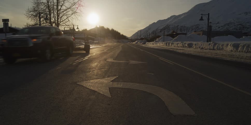 A pickup truck towing a trailer slowly drives toward the camera and turns left, following a painted arrow on the pavement of a snowy road at sunset