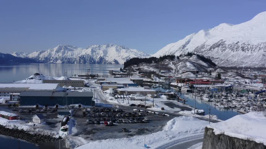 An aerial view reveals a quaint, snow-covered coastal town with a busy harbor nestled among towering mountains on a sunny winter day