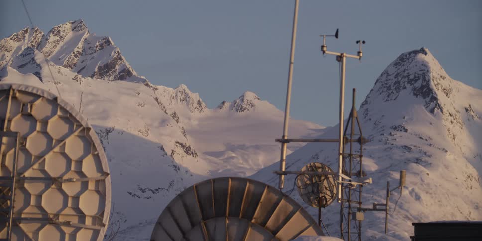 A remote weather station with satellite dishes stands before a vast, snow-covered mountain range at dawn