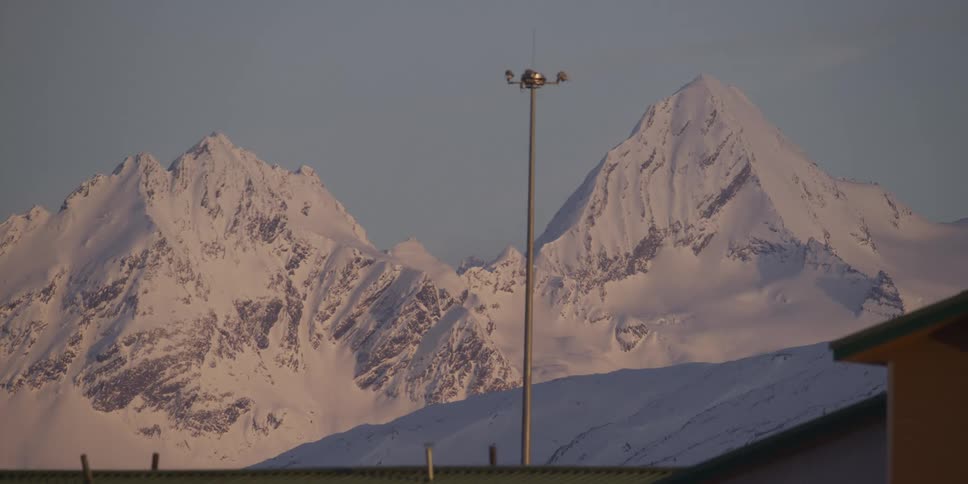 A static shot shows towering snow-covered mountains against a clear blue sky