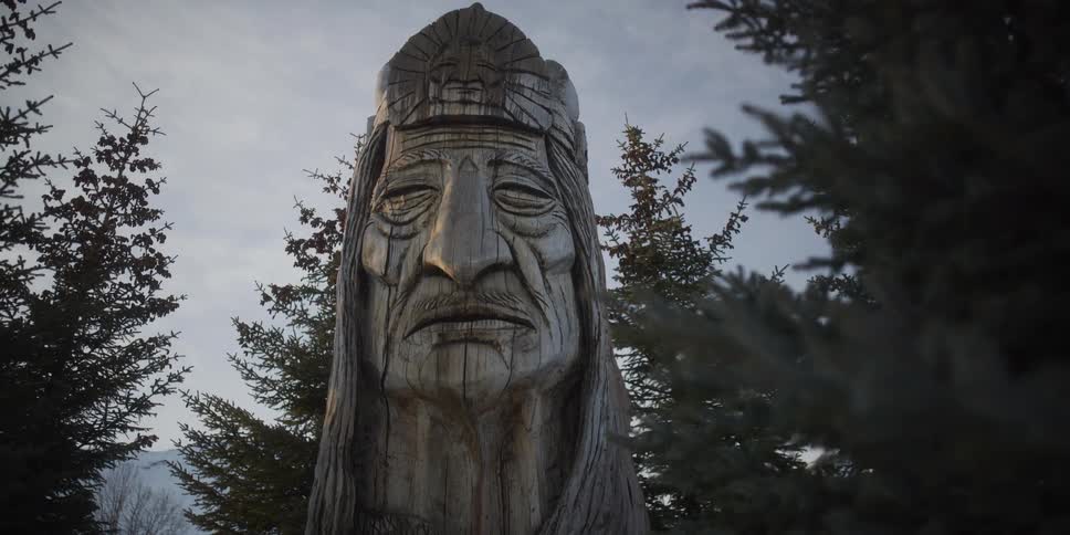 A majestic wooden carving of a First Nations chief's head stands watch over a snowy, tree-lined landscape at dusk