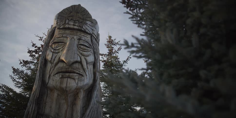 A low-angle view of a weathered wooden carving of an indigenous elder stands stoically next to evergreen trees